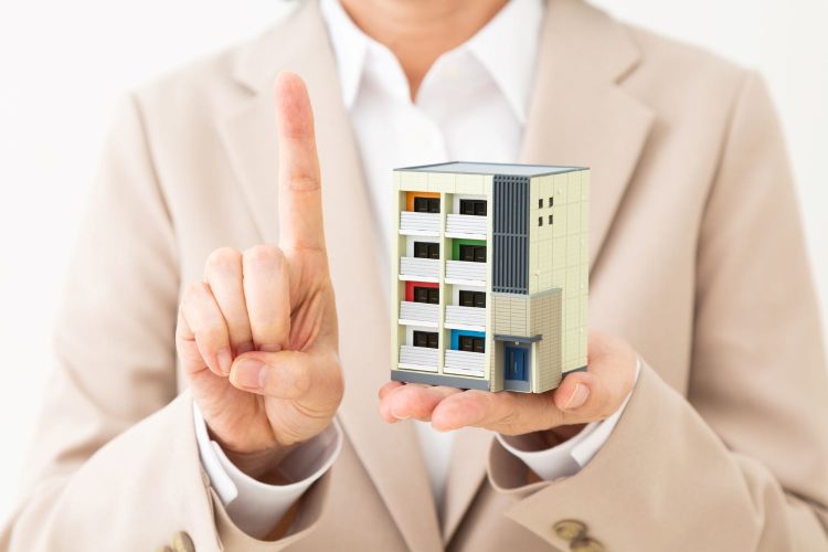 マンションの模型を持って人差し指を立てるスーツ姿の人