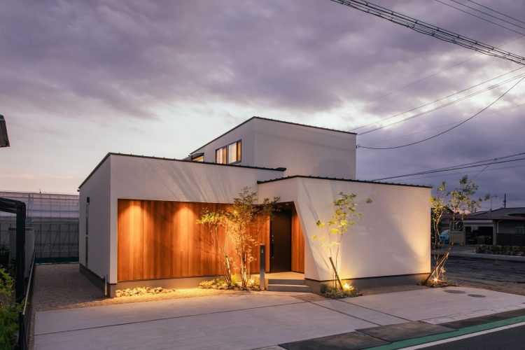 夕暮れ時にライトアップされ木目の外壁が温かく浮かび上がる白いモダンな住宅