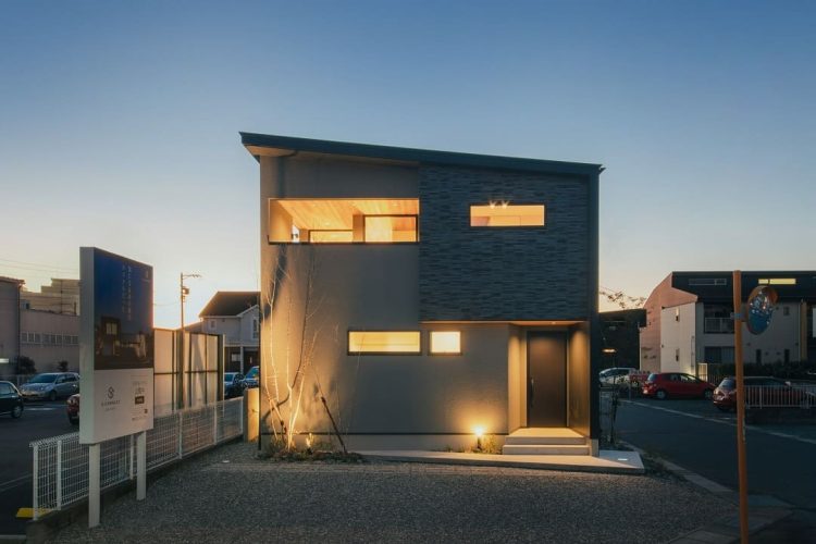 夕暮れ時に窓からの暖色の明かりが漏れるグレーの外壁の二階建て住宅