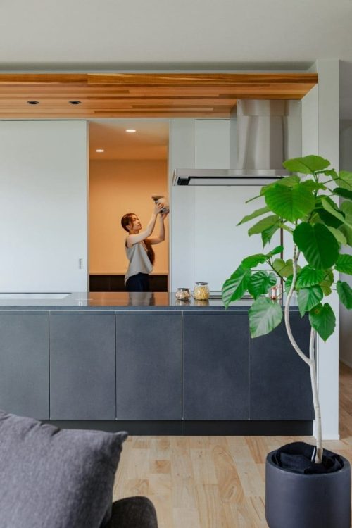 ALT_写真7_グレーのキッチンカウンターと観葉植物の奥にあるパントリーで家事をする様子